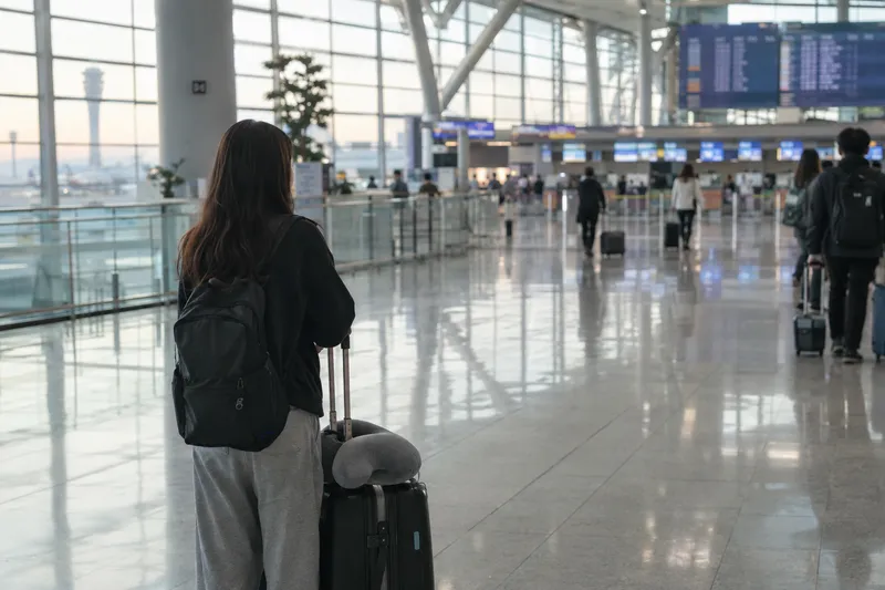 A woman with a backpack and rolling suitcase standing in an airport departure hall, looking toward the flight information boards (illustrative image)