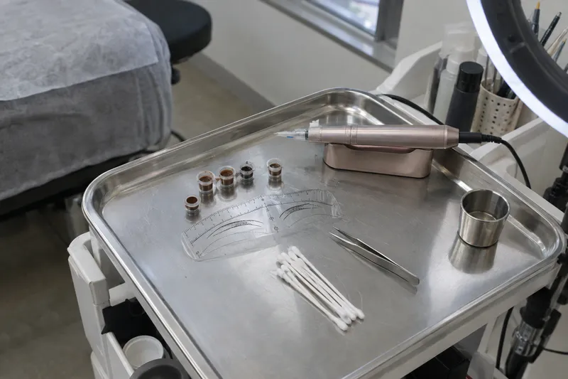 A metal tray of semi-permanent makeup tools for eyebrows, including pigment cups, a brow-mapping ruler, and a handpiece (illustrative image)