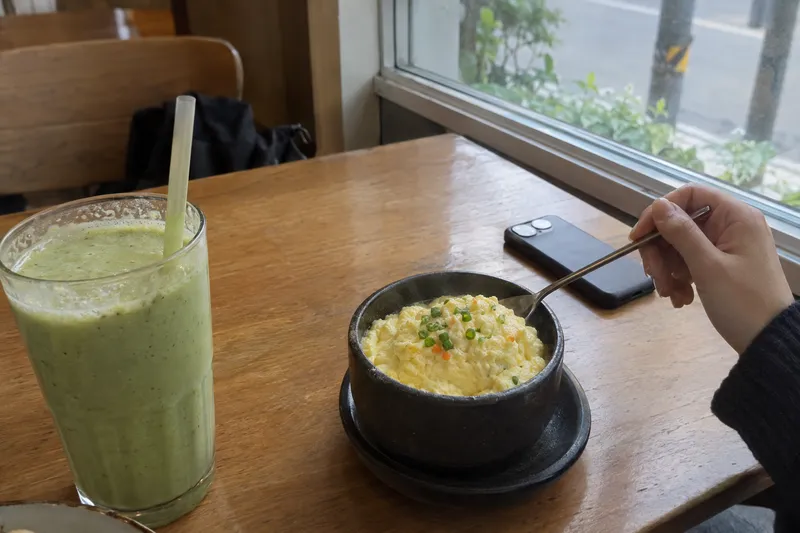 A cafe table with a green smoothie and a bowl of soft egg rice next to a phone, a simple soft-food meal (illustrative image)