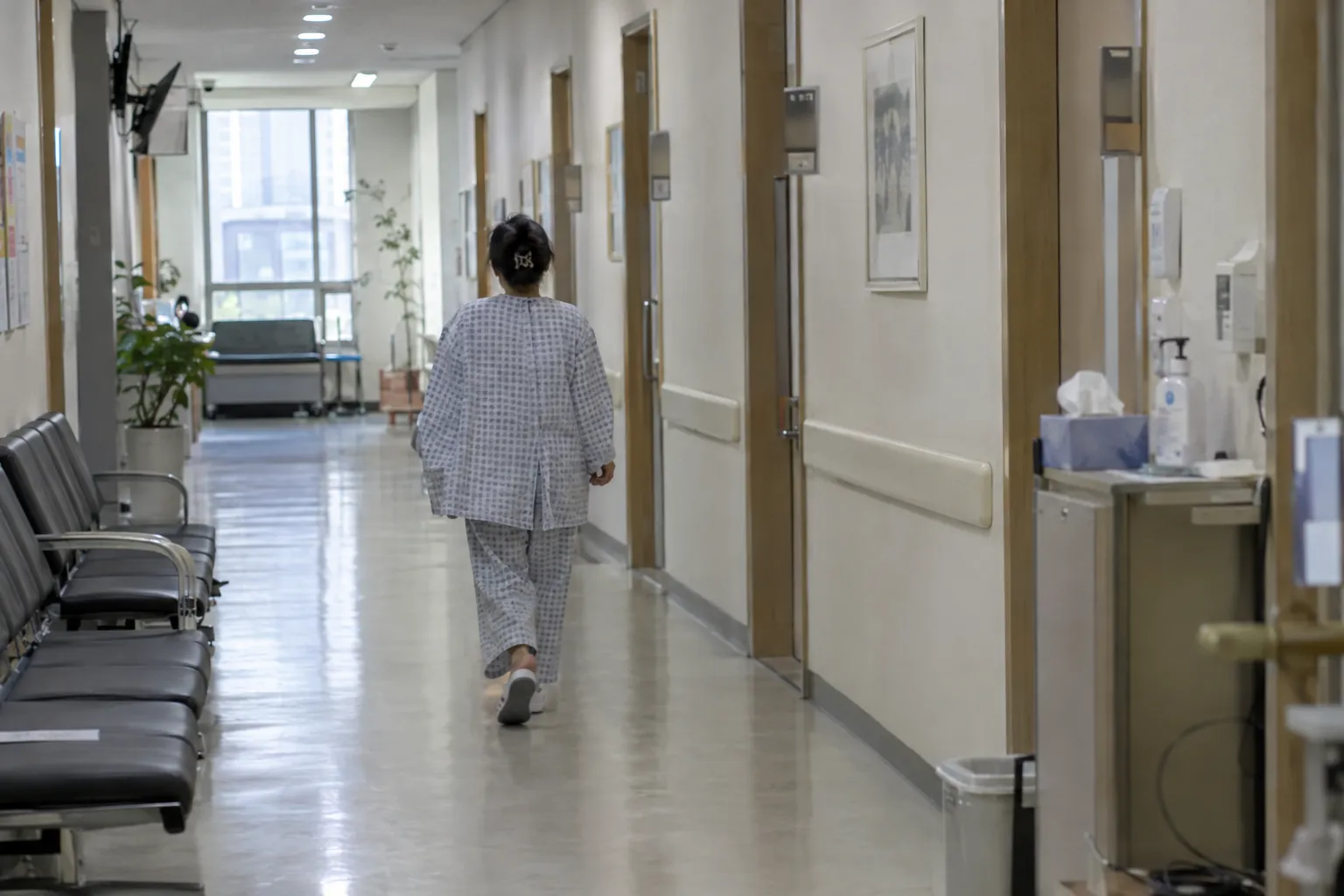 A hospital corridor with a patient in checkup pajamas walking away from the camera, past waiting chairs and treatment rooms (illustrative image)