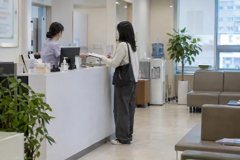 A clinic reception desk where a staff member in scrubs hands paperwork to a patient standing with a bag, seen across a waiting area with plants (illustrative image)