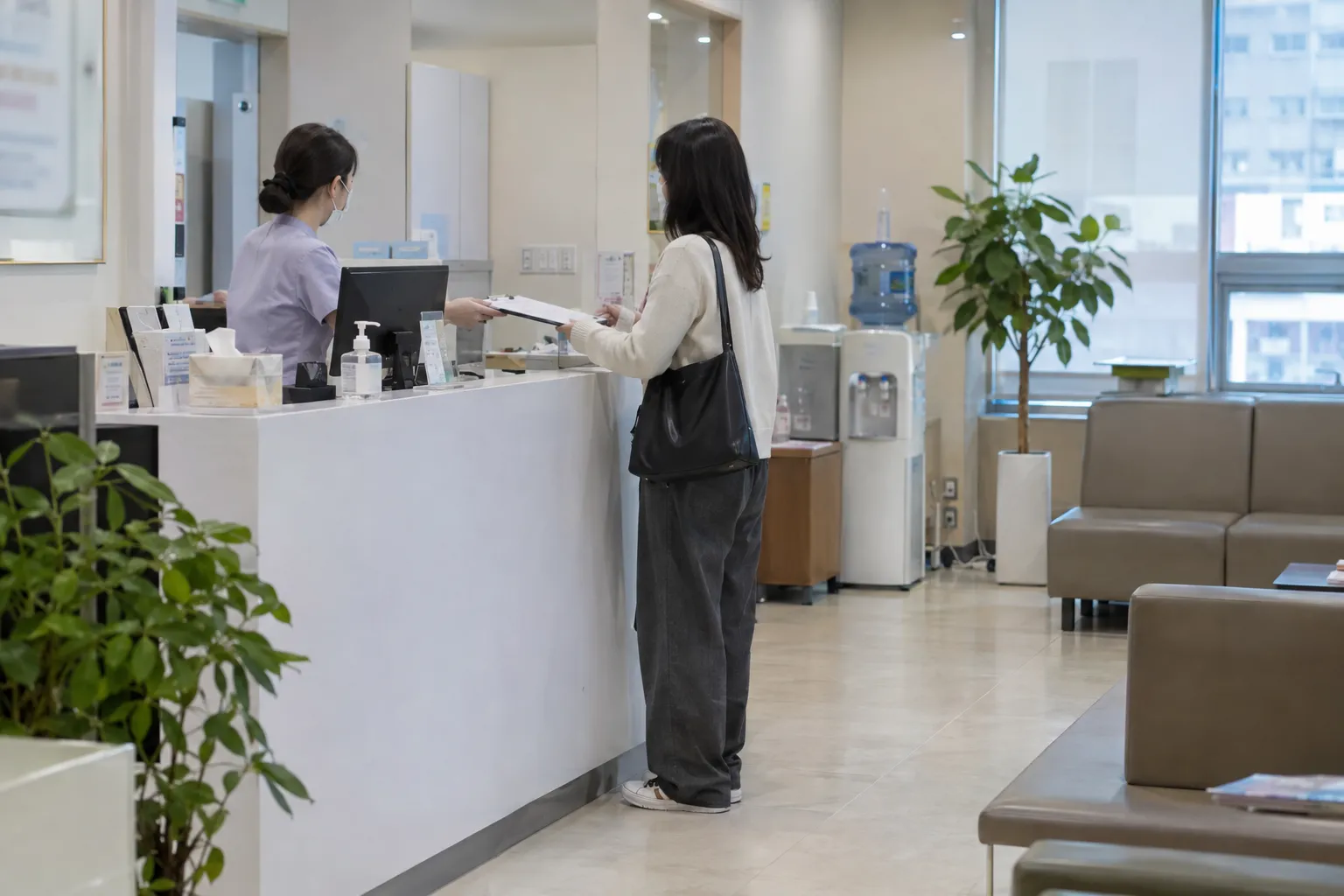A clinic reception desk where a staff member in scrubs hands paperwork to a patient standing with a bag, seen across a waiting area with plants (illustrative image)