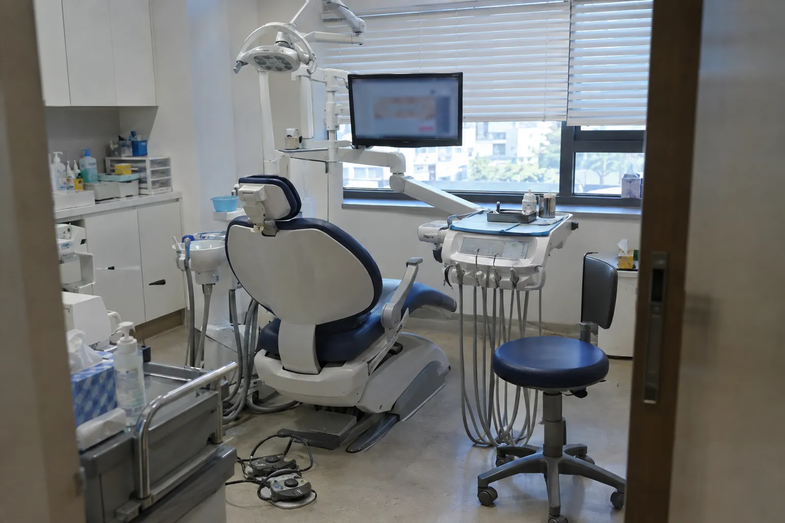 An empty dental treatment room with a reclined chair, overhead light, and a monitor beside the window (illustrative image)
