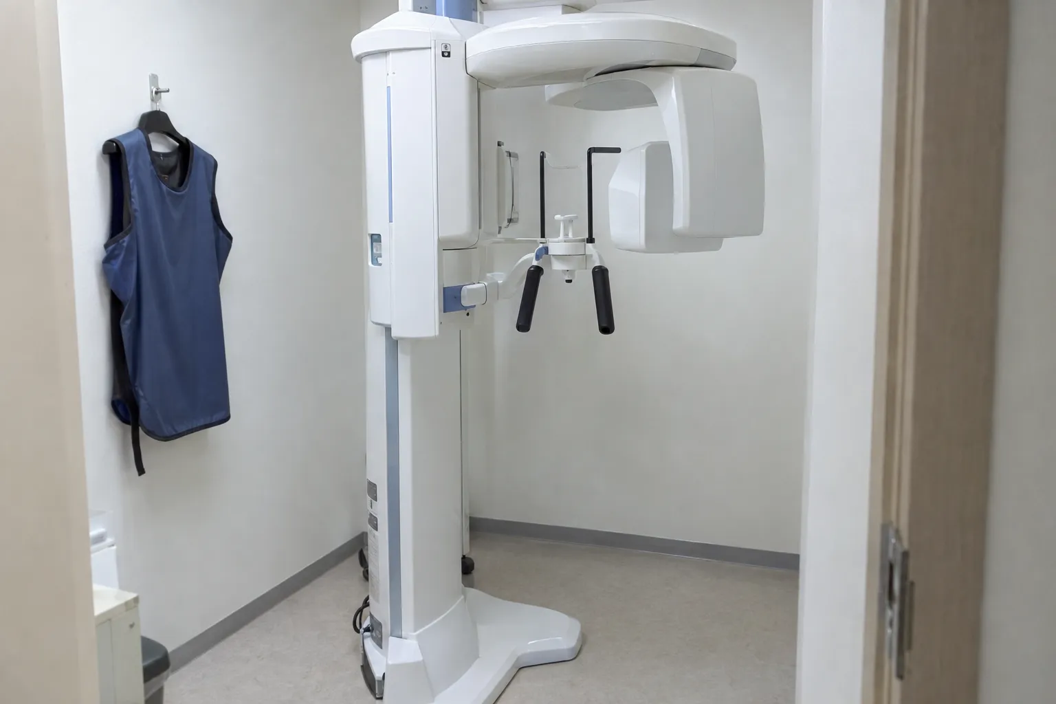 A panoramic dental X-ray machine standing in a small room, with a lead apron hanging on the wall (illustrative image)