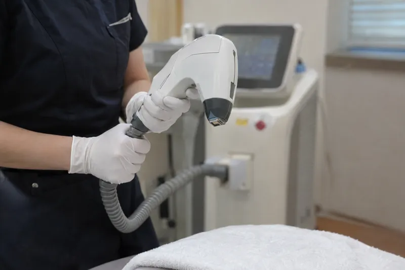 Gloved hands holding a laser treatment handpiece connected to a machine, positioned over a towel-draped treatment bed (illustrative image)