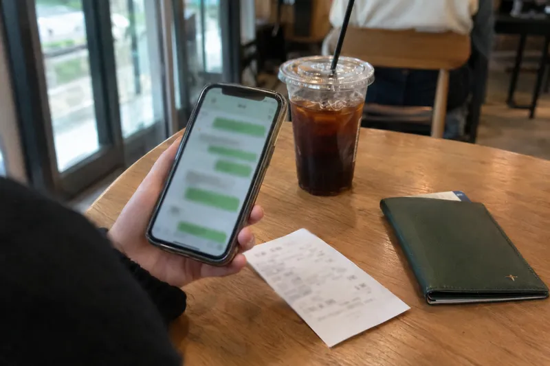 メッセージアプリの画面を開いたスマートフォンを持つ手、カフェのテーブルにはアイスコーヒーやレシート、財布が置かれている様子（イメージ画像）