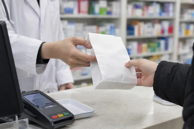 A pharmacist in a white coat handing a paper medicine bag across the counter to a customer (illustrative image)