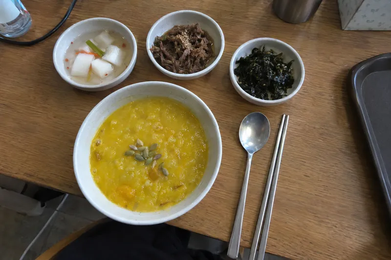 A tray of soft porridge with small side dishes and a spoon and chopsticks, a simple soft-food recovery meal (illustrative image)