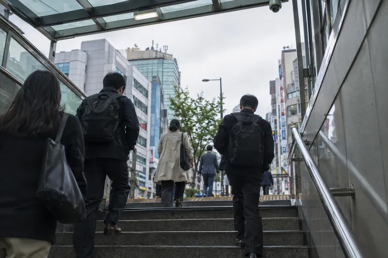 People climbing subway exit stairs toward a busy city street lined with buildings (illustrative image)