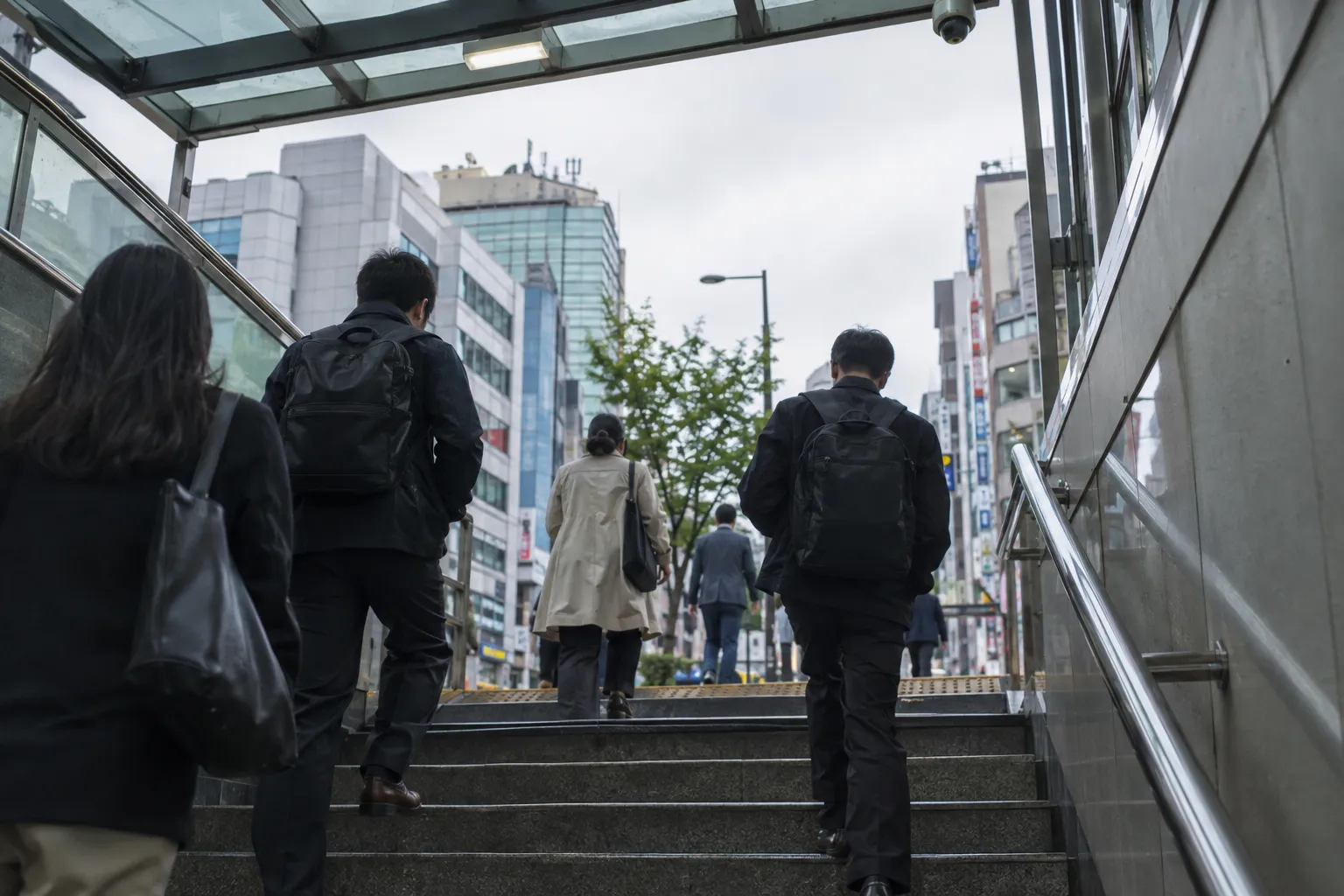 People climbing subway exit stairs toward a busy city street lined with buildings (illustrative image)